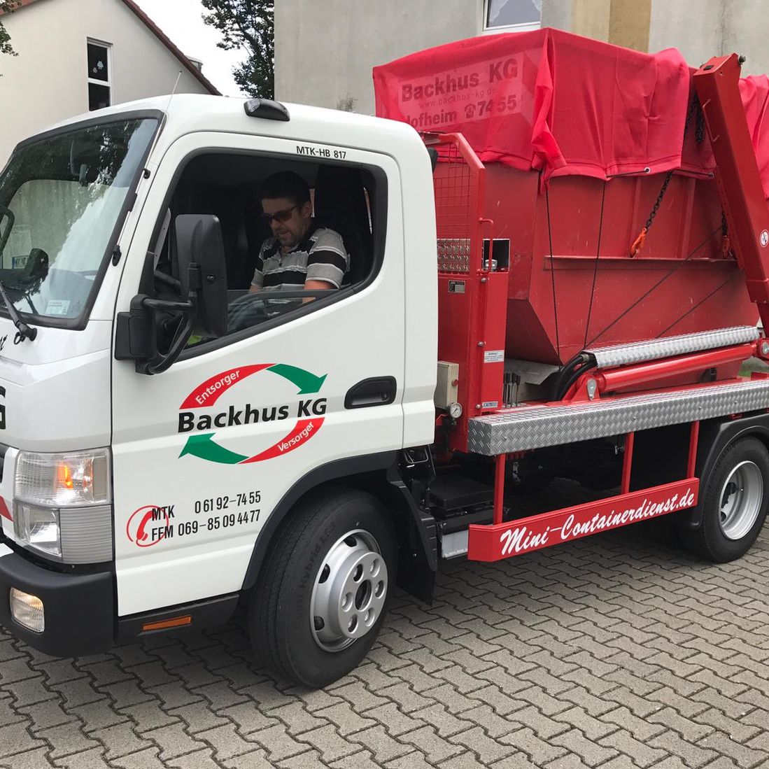 Ein Fahrer startet mit einem Mini LKW, der mit einem roten, mit einer roten Plane abgedecktem Container.
